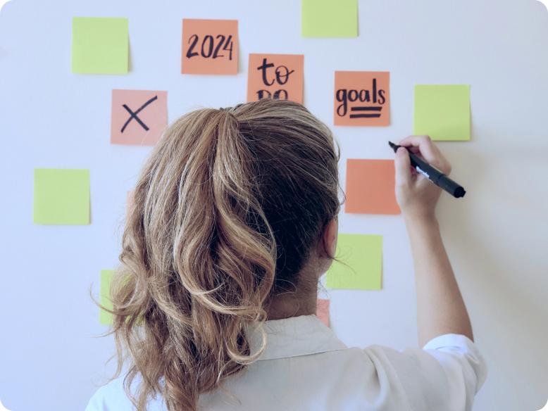 Eine Person steht vor einer Wand und schreibt mit einem schwarzen Marker auf bunte Klebezettel. Auf den Zetteln sind Begriffe wie „20%“, „to go“, und „goals“ zu sehen.