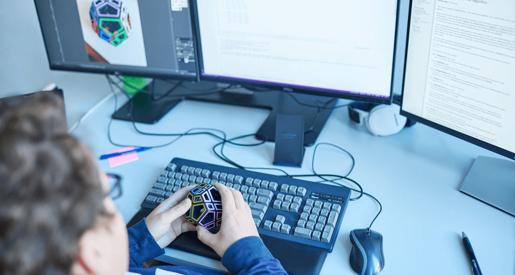 Arbeitsplatz mit Tastatur, Monitoren und einem Zauberwürfel in der Hand